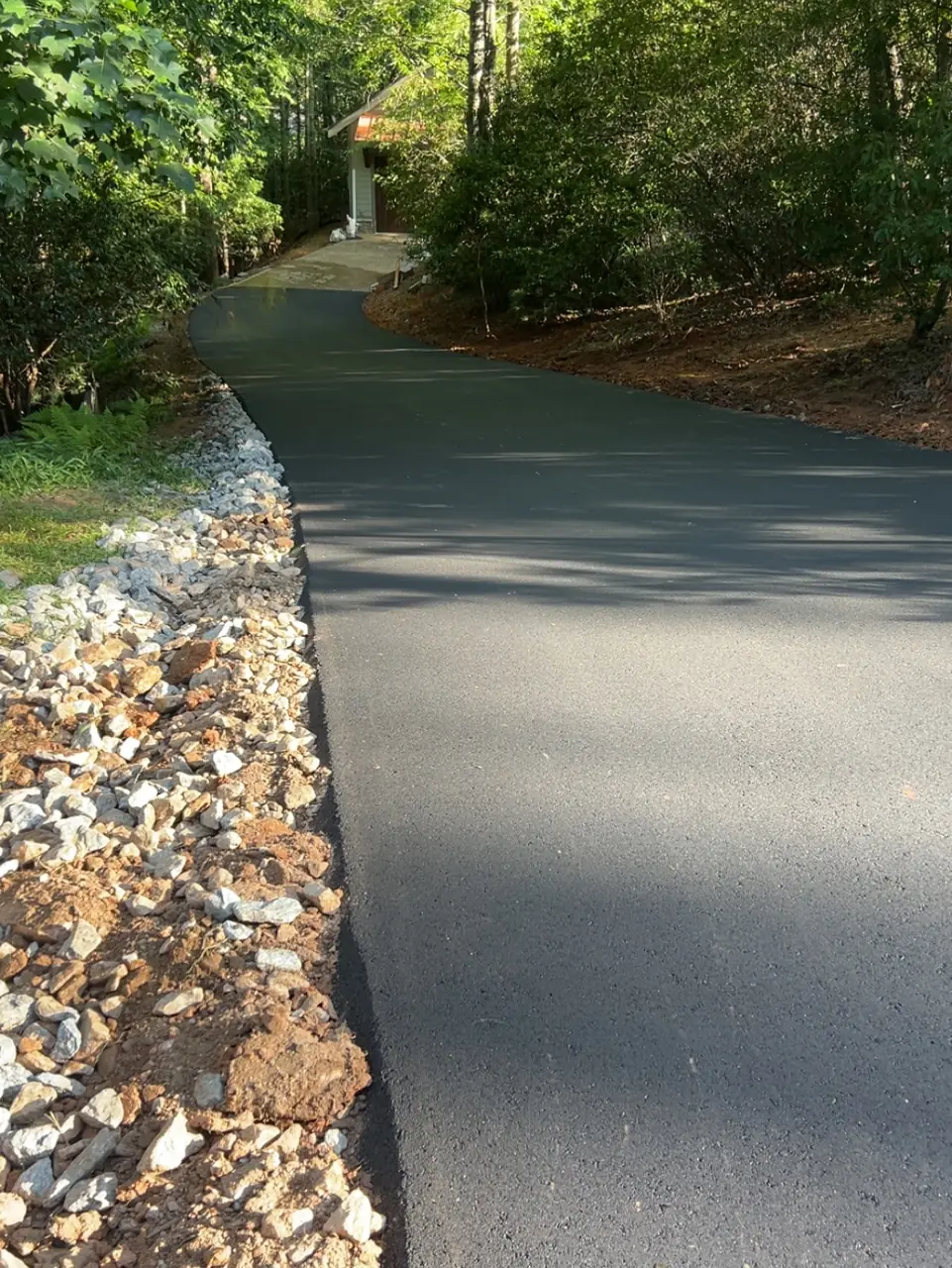 A narrow paved lane with a stone-lined edge on a wooded lot